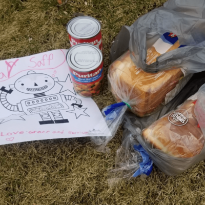 Two packages of sliced white bread, two cans of soup and a child's drawing of a robot are on grass.