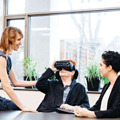 Two young women are sitting on either side of a senior aged woman wearing a virtual reality headset.